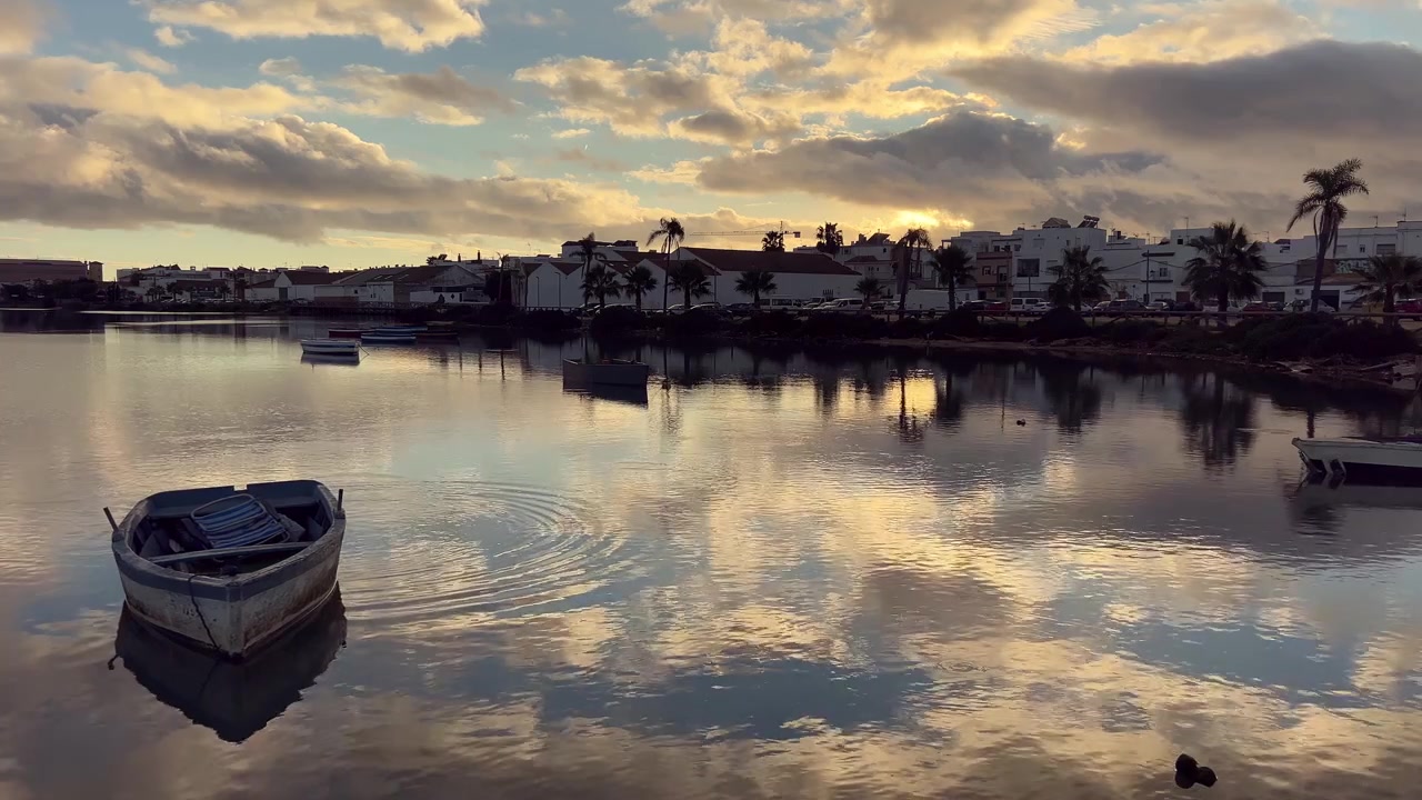 Pequeno barco de pesca de madeira flutuando em águas calmas, refletindo o belo céu do pôr do sol e a paisagem urbana de Barbate, uma cidade encantadora na Andaluzia, Espanha, criando uma vista tranquila e pitoresca.