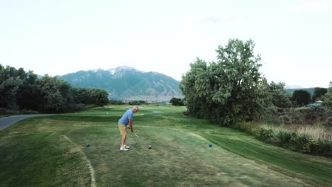 Disparo-De-Drones-Volando-Hacia-Un-Golfista-En-Un-Tee-Box-Que-Balancea-Y-Conduce-Su-Pelota-De-Golf-Hacia-La-Calle