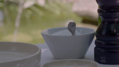 close-up shot of a dining setup in provence, france, featuring a small white bowl of salt with a spoon, a black pepper grinder, and blurred water feature in the background.