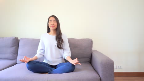 dressed in jeans and a sweater, a young woman with closed eyes strikes a yoga pose on a couch