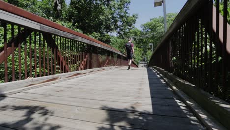 Young-Man-With-A-Backpack-And-A-Camera-Walking-On-A-Wooden-Path-Surrounded-By-Green-Trees-On-A-Sunny-Day---Low-Level-Shot