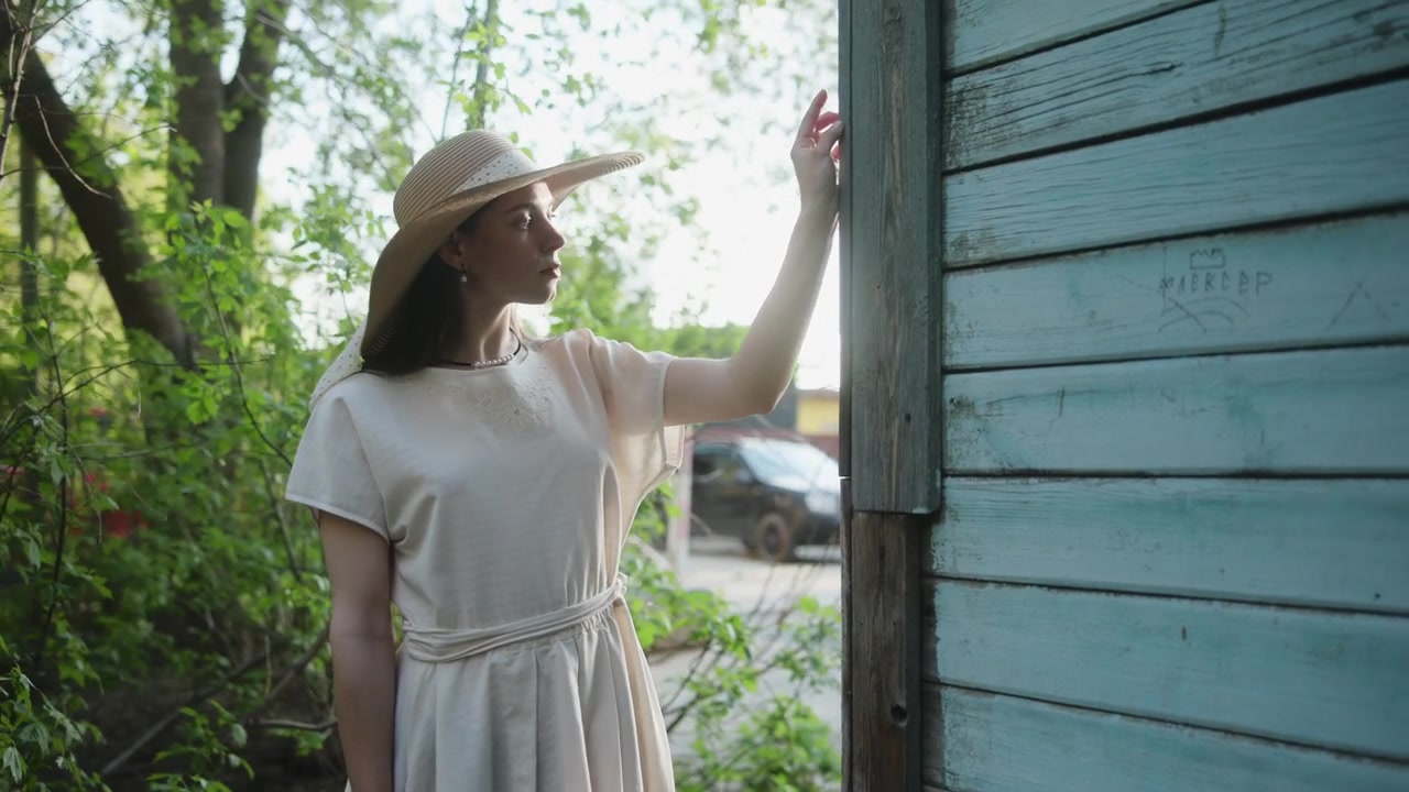 Una giovane donna che indossa un vestito leggero e un cappello sta in piedi accanto a un muro di legno blu invecchiato, protendendosi verso l'ingresso in un ambiente esterno luminoso con alberi.