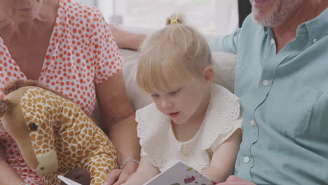 Cerca-De-Abuelos-Sentados-En-El-Sofá-Con-Su-Nieta-En-Casa-Leyendo-Un-Libro-Juntos