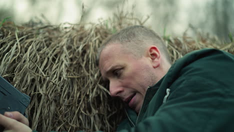 a close view of a man in a green jacket lying in a ditch against dry grass with a pistol in hand, the man looks exhausted and tense