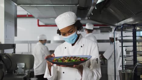 Portrait-of-african-american-female-chef-wearing-face-mask-presenting-dish