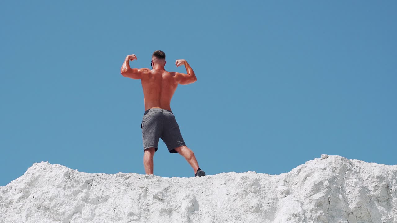Bodybuilder athlete posing with muscles in nature against a blue sky. A man stands with his back