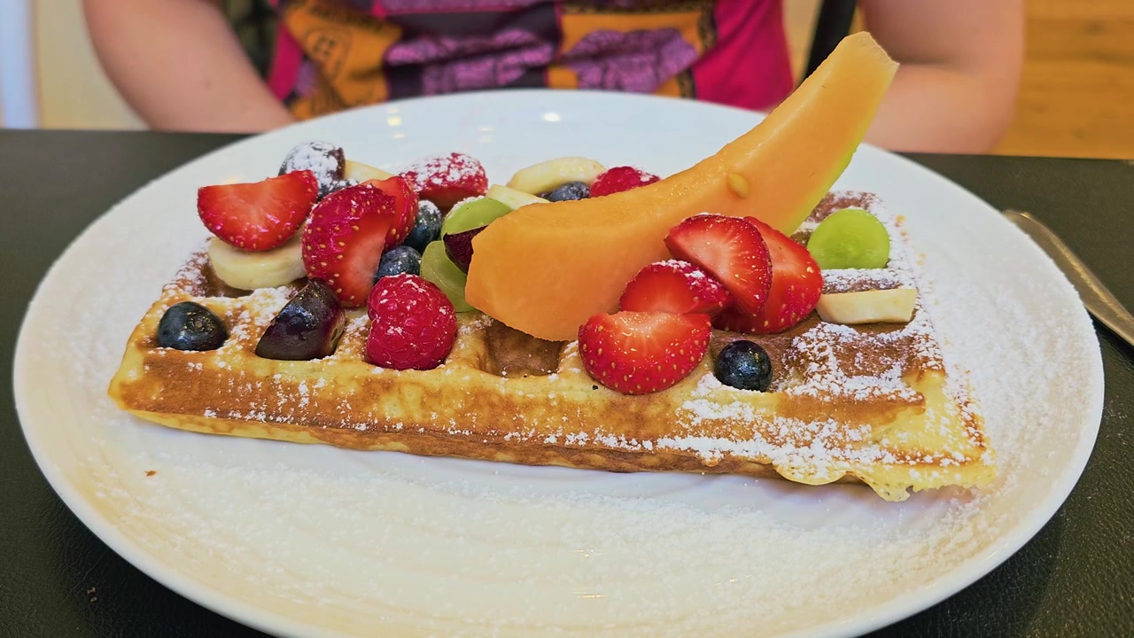 Primo piano di un dessert waffle dorato coperto con frutta fresca mista, fragole a fette, melone, banana, frutti di bosco, uva e zucchero a velo su un tavolo da caffè durante il servizio del brunch