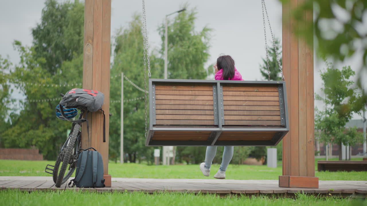 Žena na houpačce vedle kola, batoh opřený poblíž, zamračený park, jemný pohyb houpání, klidná zamyšlená nálada, zeleň a stromy rámující scénu, filmová malá hloubka ostrosti, městský únik, zamyšlená
