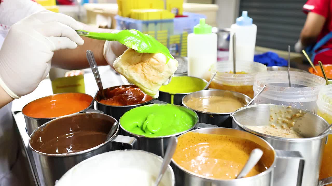 Gloved hands spread vibrant sweet sauces on bread at a busy Bangkok street food stall