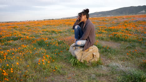 una linda chica llorando y luciendo emocional y triste sentada sola en un campo de flores silvestres con el viento soplando su cabello al atardecer a cámara lenta