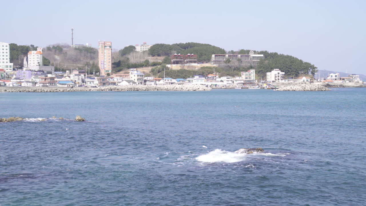 Sea waves hitting onto a small rock with a view of a small port at the background (shoot info: Cin2, 1080p, 59.94fps)