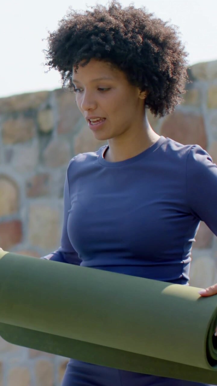 Video verticale: Donna con top blu scuro che cambia presa, srotola un tappetino verde contro un muro di pietra, mentre un tappetino scuro passa.