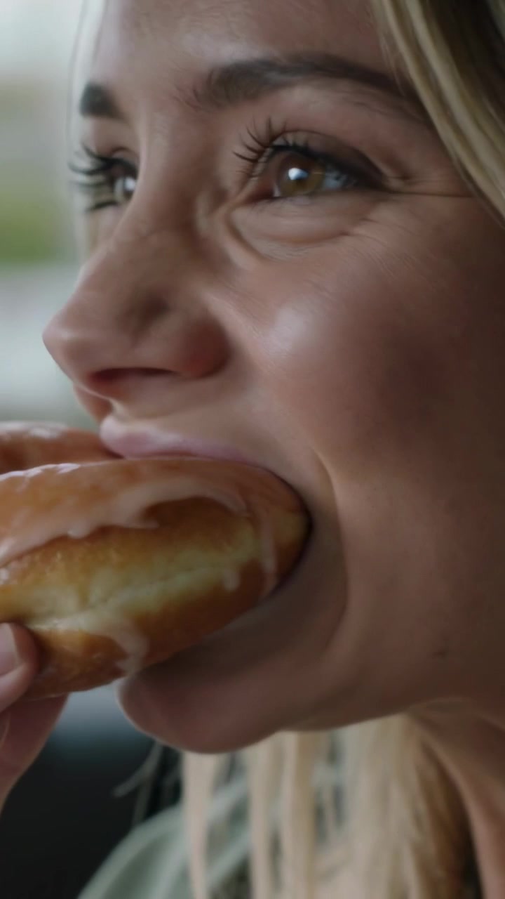 Vertical video: Raising glazed donut, woman in green jacket taking bite while snacking in car