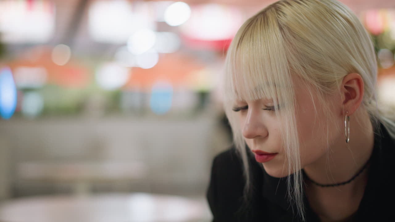 Close up of blonde lady with bangs applying lipstick delicately indoors, wearing hoop earrings and black choker, showing beauty routine, elegance, and stylish lifestyle with blurred warm background
