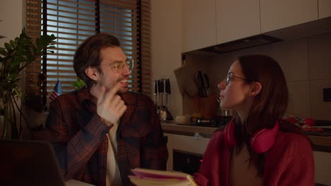 a confident young brunette girl with glasses in a red sweater and wireless headphones studies foreign languages together with her brunette male teacher who teaches the girl the correct pronunciation in a modern apartment in the kitchen