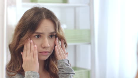 upset woman looking face and touching skin front bathroom mirror in home