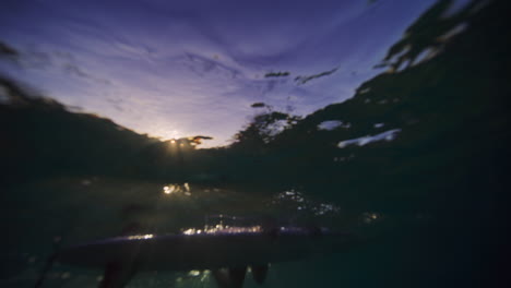 Underwater-below-surface-view-of-girl-hanging-off-edge-of-longboard-at-sunrise