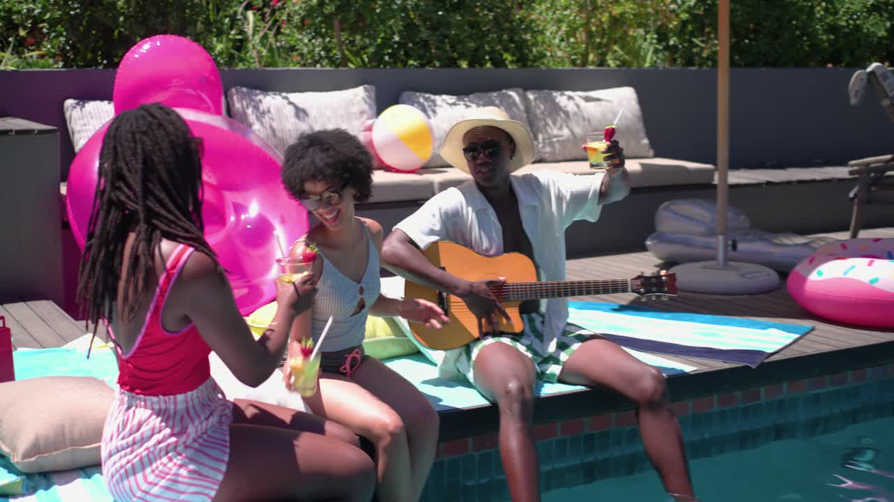 Amici afroamericani seduti a bordo piscina mentre un uomo strimpella la chitarra e una donna porge bevande fredde.