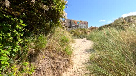 point-of-view walk along a sandy, grassy path toward a graffiti-covered world war-era fort under bright daylight in coastal dunes near dunkirk, france