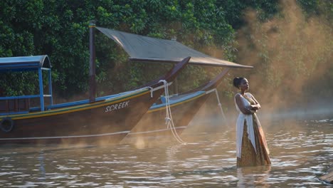woman standing in calm water, dressed in traditional clothing, with arms crossed near two canopied wooden boats, surrounded by mist and lush vegetation at sunrise