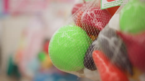 hand view of woman touching rubber children toys, pressing round green ball in mesh packaging, exploring texture and shape, colorful curved play items blurred in background in retail environment