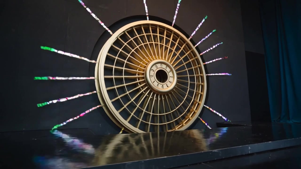 Strisce LED che si illuminano, ruota dorata che ruota in senso orario e si sposta a destra sul palco, creando fasci di luce.