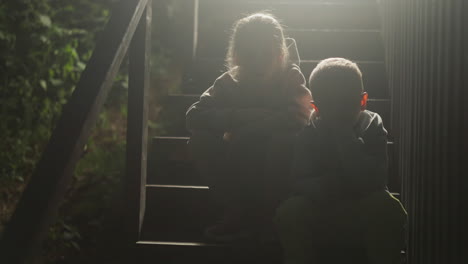 children silhouettes on cottage stairs at night. little boy and girl rest sitting on steps at country house. calm kids spend time together in evening