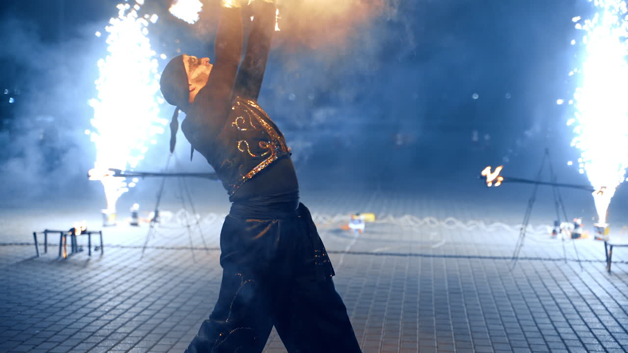 Man dancing fearlessly with burning object. Artist rotates burning stick at night. Fireworks at backdrop.