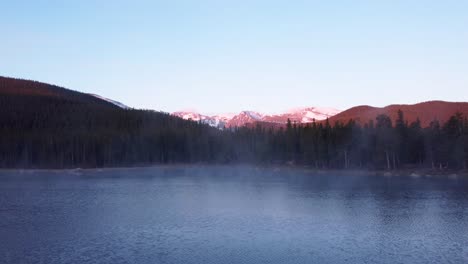 Alpenglow-on-snowcapped-Rocky-Mountains-with-steaming-lake-during-the-sunrise,-aerial