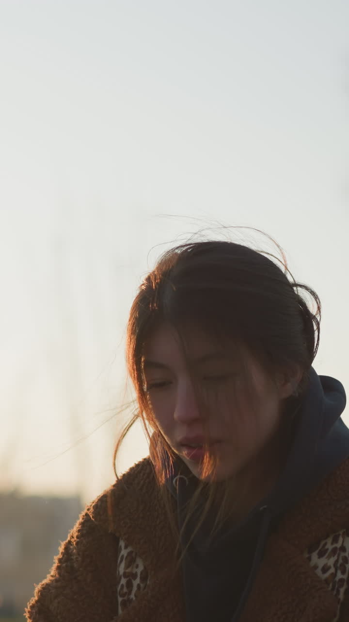 한 우울한 소녀가 일몰에 공원을 걸어가며 머리카락이 얼굴을 부분적으로 고, 갈색 코트와 후디를 입고, 그녀는 생각에 빠진 것처럼 보이며, 부드럽고 희미한 빛으로