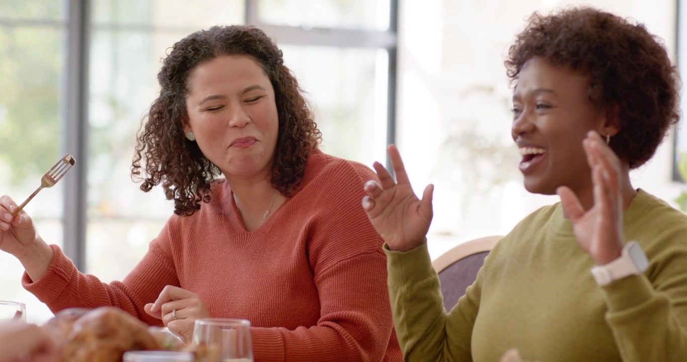 Diverse female friends sharing meal at dining table by bright window, left woman tasting roast