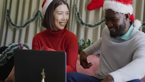 video of happy diverse couple in santa hats smiling and making christmas laptop video call at home