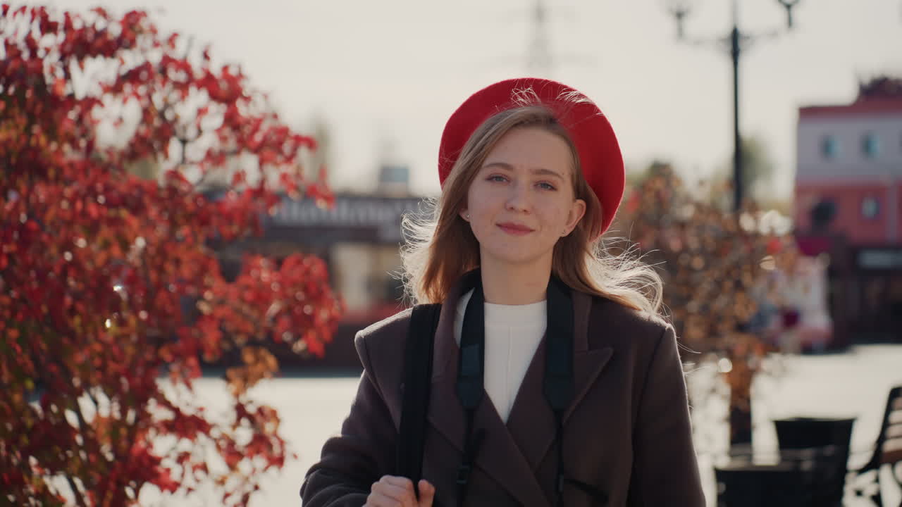 Giovane donna con berretto rosso che cammina attraverso una piazza illuminata dal sole con fogliame rosso, portando una borsa tote e un cappotto, un sorriso disinvolto, panchine e lampioni, un ritratto di strada spontaneo con calda retroilluminazione e un'accogliente atmosfera autunnale.