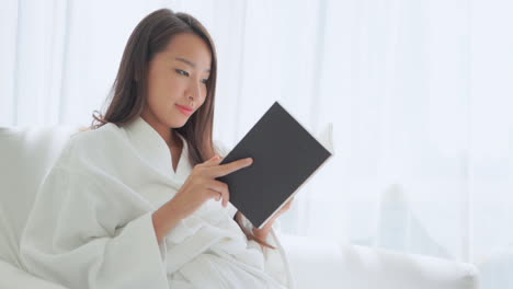 smiling pretty asian woman lying on the hotel white bed wearing a white dressing gown and reading a book in the morning daytime