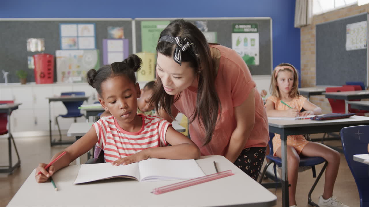 feliz maestra diversa ayudando a una colegiala en el escritorio en la clase de escuela primaria, cámara lenta