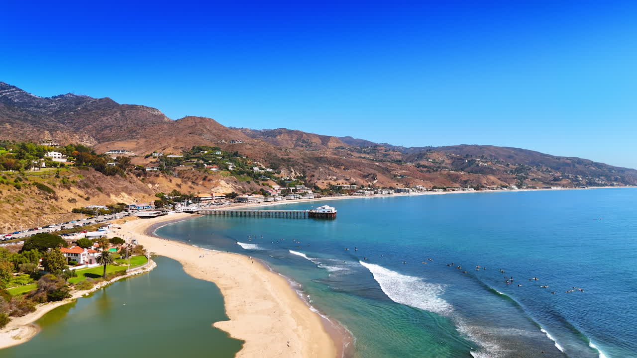 Multiple surfers float on the waves of the Pacific Ocean in Malibu. Pier on the beach of sunny Malibu, California, USA. Bare rocks at the backdrop