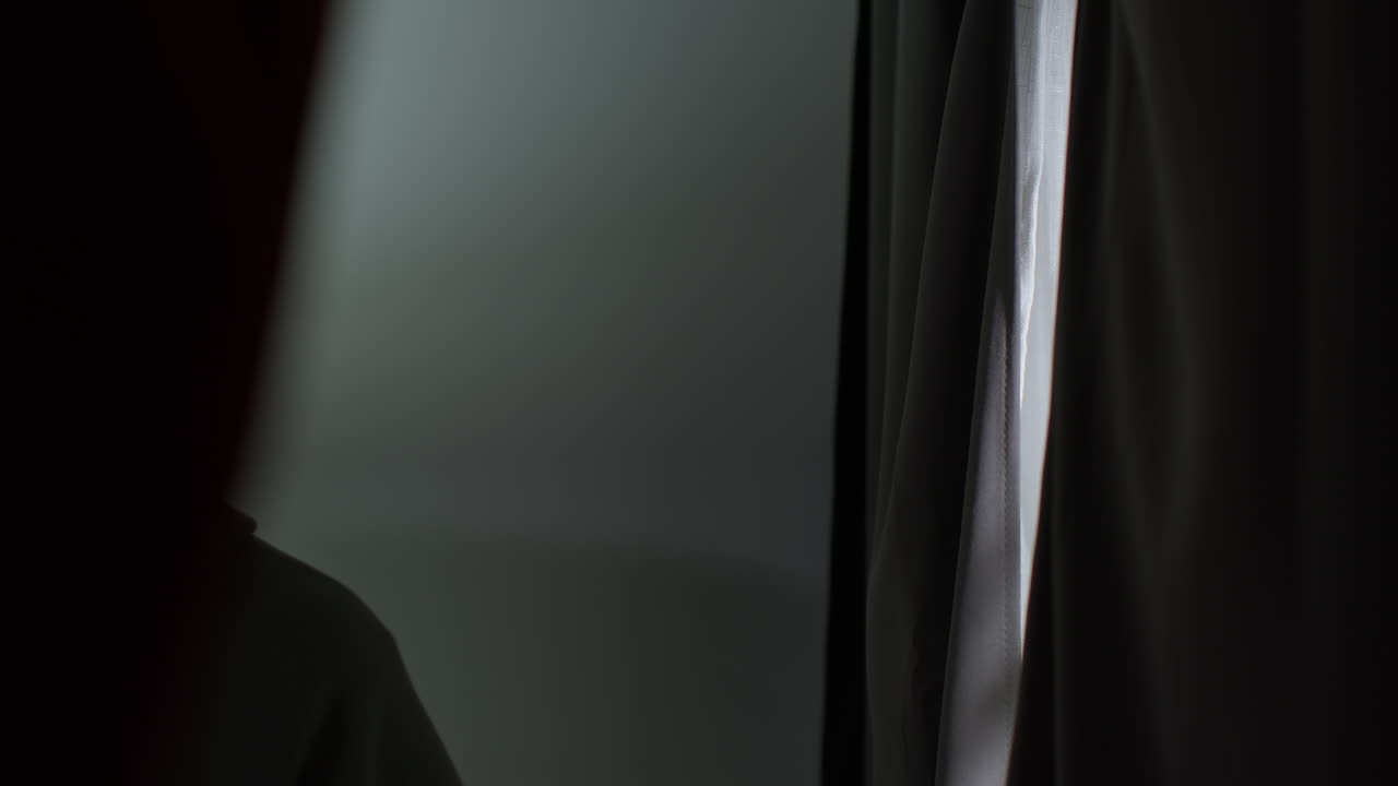 불행하고 우울한 젊은이가 집에서 커튼을 열고 창밖을 바라보고 있습니다.