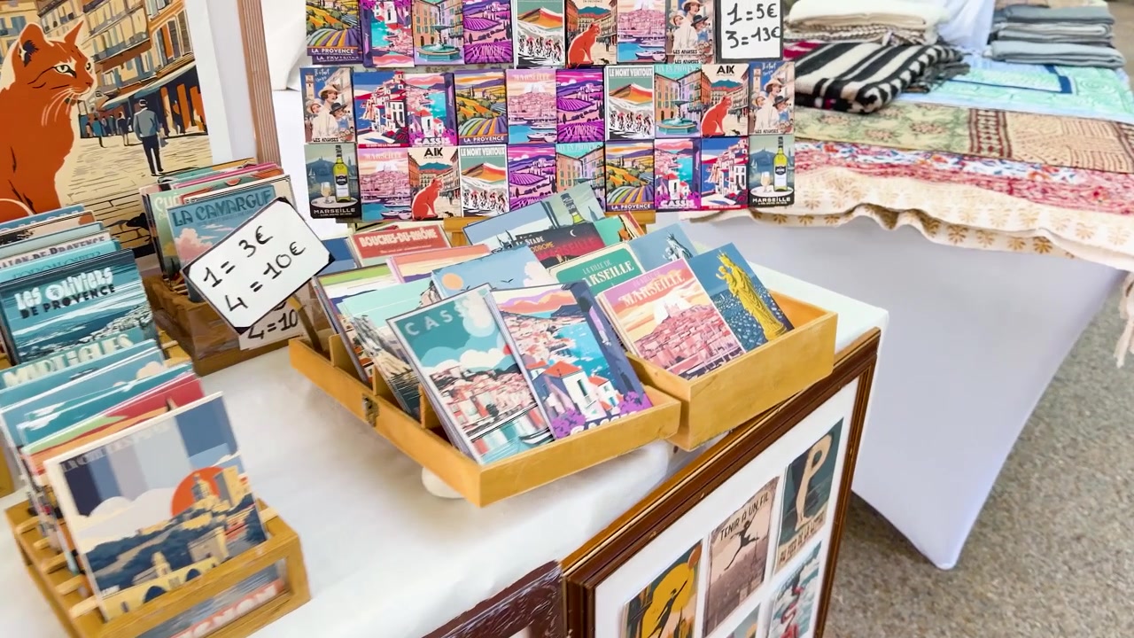 Cámara en mano panea por un puesto de mercado que exhibe postales de estilo vintage, láminas de arte y textiles.