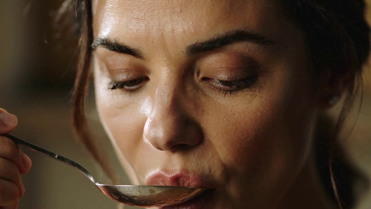 Mulher segurando uma colher de metal perto dos lábios, fechando os olhos e saboreando enquanto avalia o sabor na cozinha.