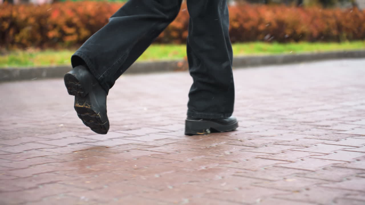 Close-up shot of human legs running during light snowfall, wearing black jeans and black boots. Background features autumn trees with leaves and wintery atmosphere, creating a serene feel