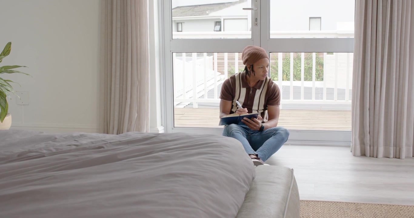 African American man sitting by door, writing in notepad pausing at balcony motion, copy space
