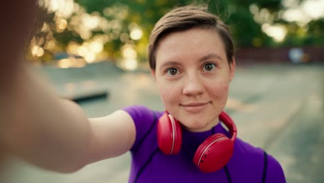 Vista-En-Primera-Persona:-Una-Chica-Con-Un-Corte-De-Pelo-Corto-Y-Un-Top-Morado-Con-Audífonos-Rojos-Se-Toma-Una-Selfie-En-Un-Parque-De-Patinaje-En-Verano