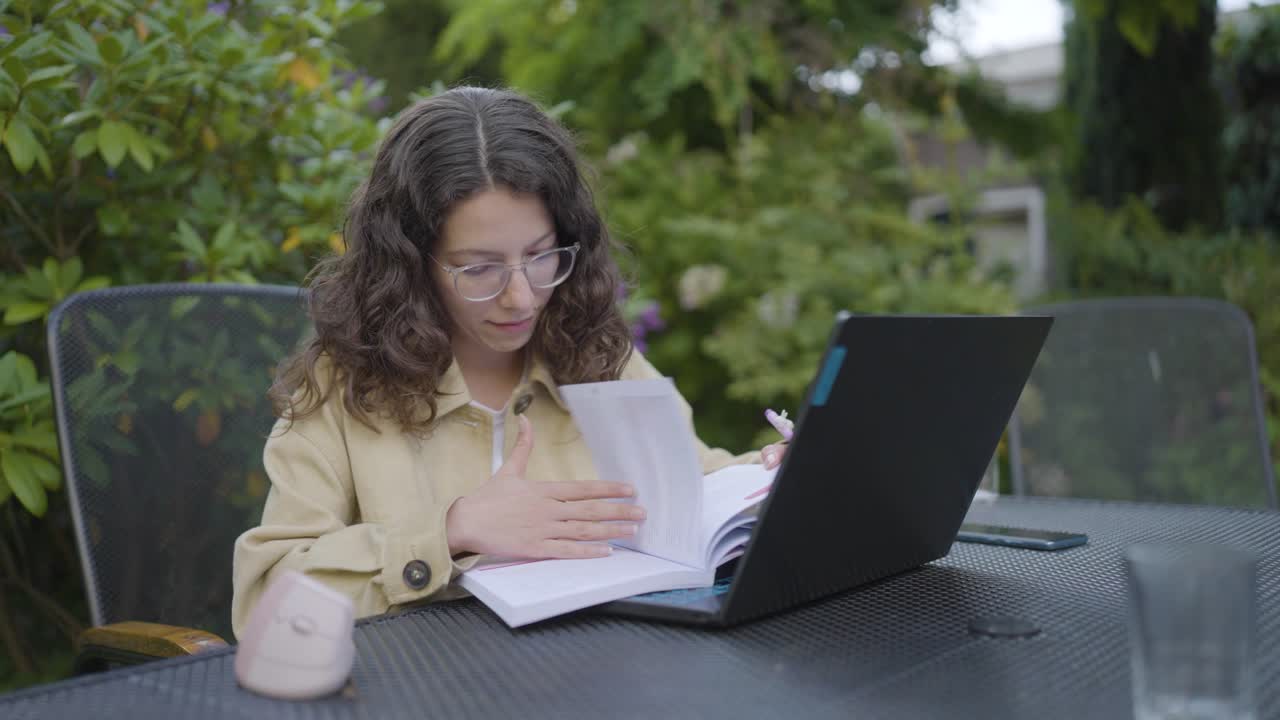 Ung kvinne som jobber på en bærbar datamaskin og leser notater i en hage, som representerer fjernarbeid og utendørs produktivitet