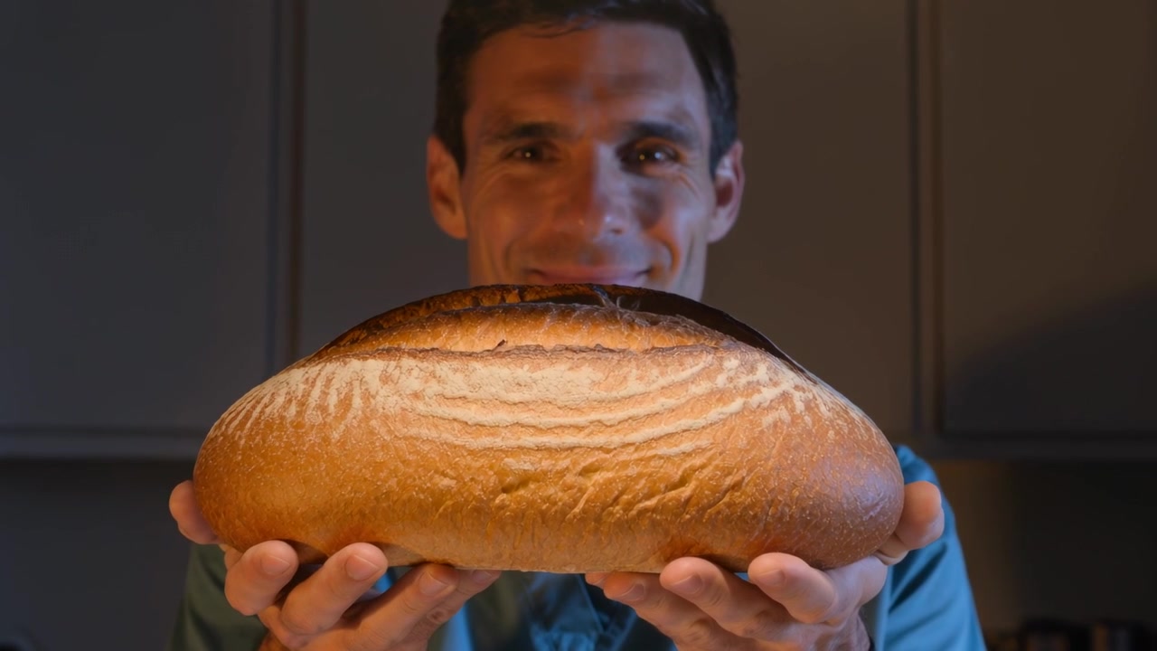 Man wearing teal-blue shirt presenting large loaf in kitchen, with recording starting to show scale