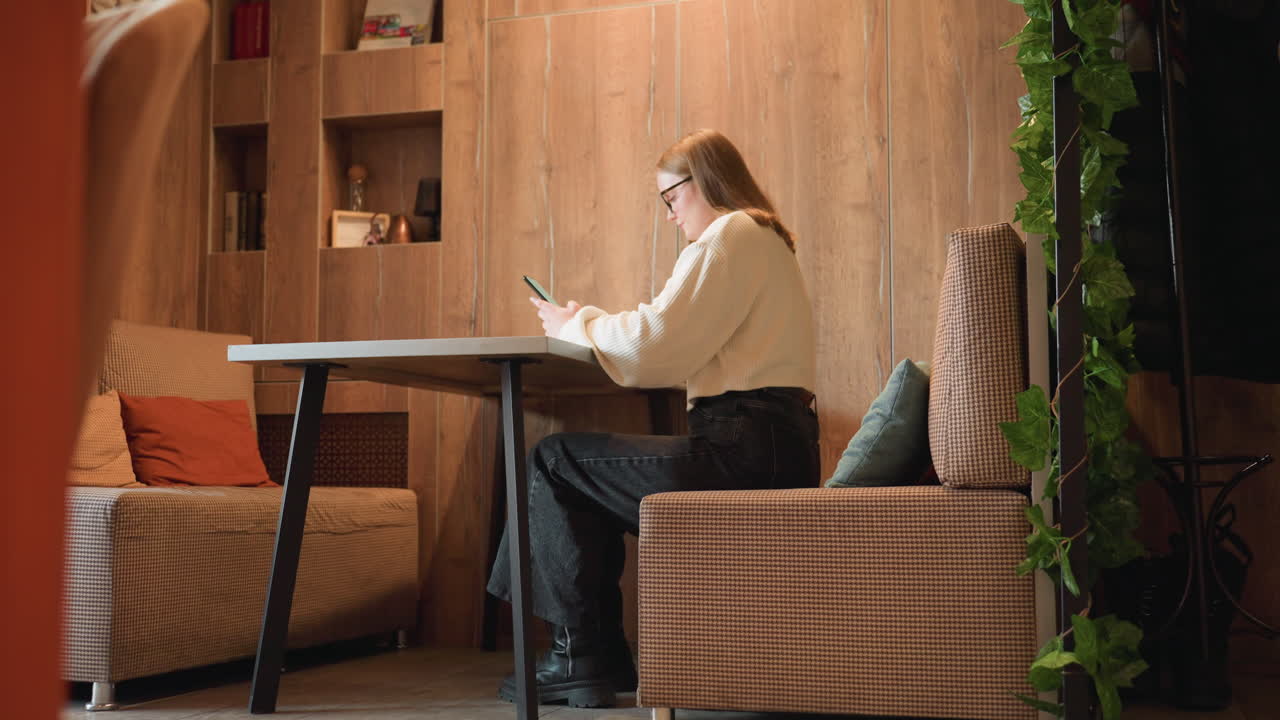 Remote worker seated in cafe responding to client message on smartphone, with empty cushion placed in front and decorative green leaves behind her, surrounded by wooden panels