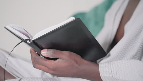 close-up of man's hand in white bathrobe holding black notebook, turning pages with focus and concentration, soft lighting highlighting notebook details, relaxed indoor atmosphere