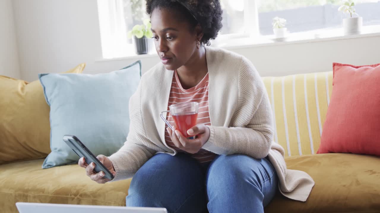 Focused african american woman working at home having tea, using smartphone and laptop, slow motion
