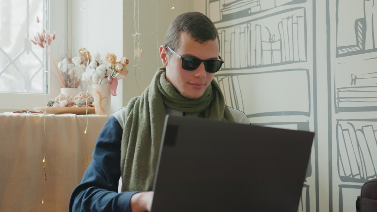 Caucasian man working on laptop in cozy home library, wearing green scarf and sunglasses, typing and researching with notebook and coffee, remote developer and writer vibe, soft daylight