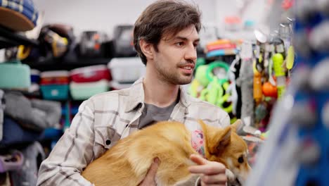 A-confident-brunette-guy-in-a-white-shirt-holds-a-red-corgi-dog-in-his-hands-and-chooses-food-for-it-in-a-pet-store