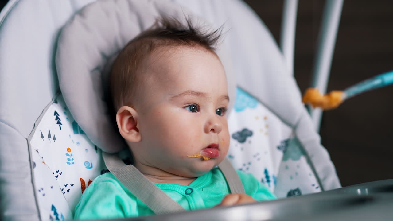 Feeding a baby sitting in a chair. Cute kid opens mouth to a spoon. Mother's giving meals to a son.